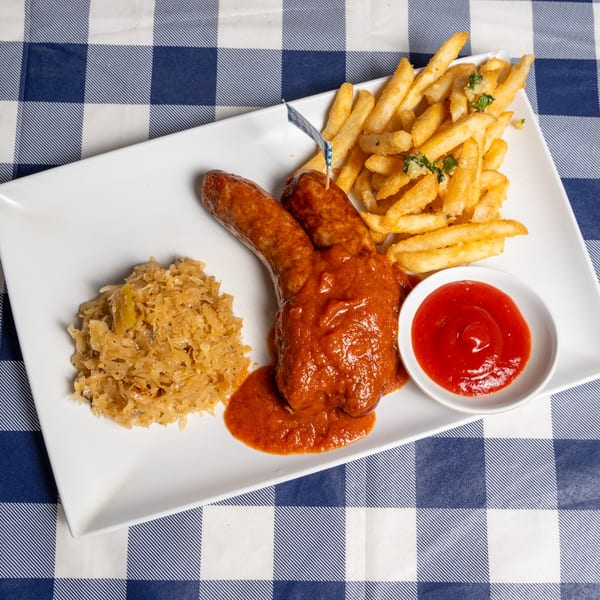Currywurst with red sauce, a side of sauerkraut, french fries, and ketchup on a white plate.