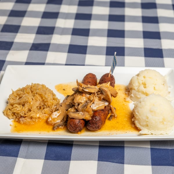 Sausages with mushroom gravy, sauerkraut, and mashed potatoes on a white plate over a blue and white checkered tablecloth.