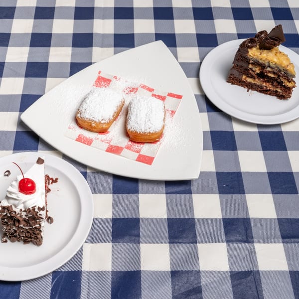 Three dessert plates on a blue and white checkered tablecloth, featuring beignets and two cake slices.