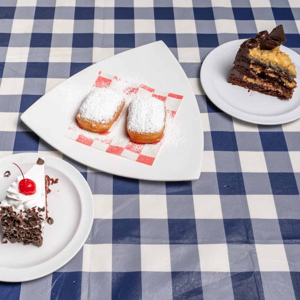 Three dessert plates on a blue and white checkered tablecloth, featuring beignets and two cake slices.