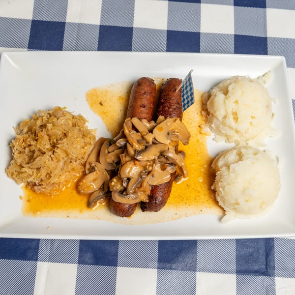 Two sausages topped with mushrooms, sauerkraut, and mashed potatoes on a white plate.