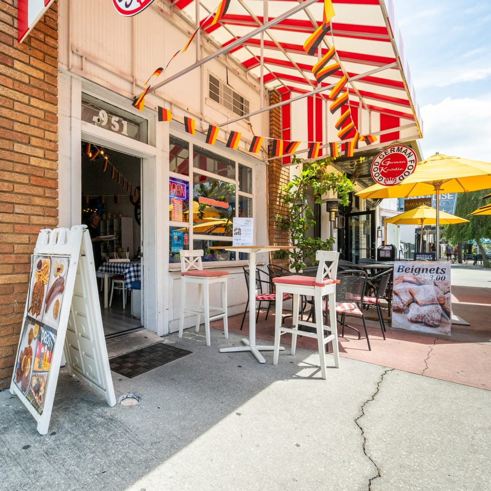 The exterior of a German restaurant with a striped awning, outdoor seating, and German flags.