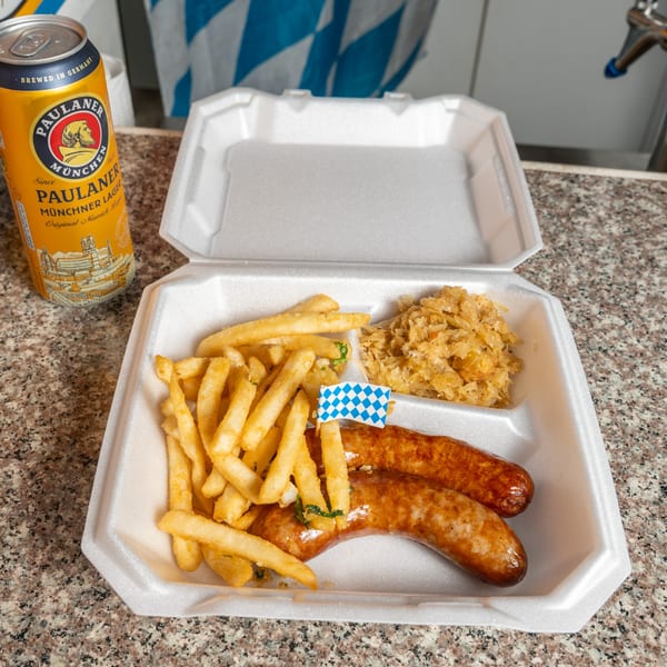 A Paulaner beer can next to a takeout container with two sausages, fries, and sauerkraut.