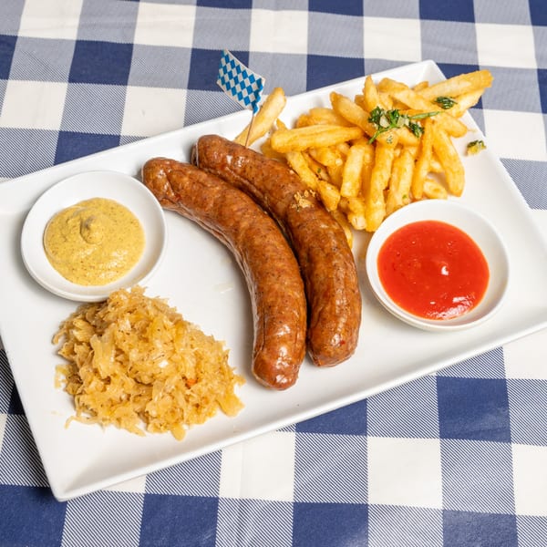 Two sausages, fries, sauerkraut, mustard, and ketchup on a white plate over a blue and white checkered tablecloth.