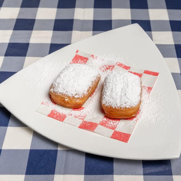 Two powdered sugar beignets on a white plate with red and white checkered paper, on a blue and white checkered tablecloth.