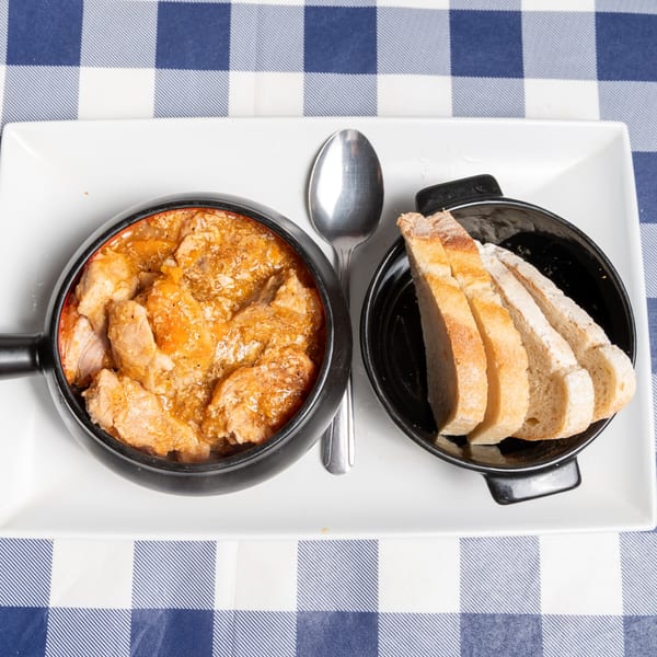 Chicken stew in a black pot with sliced bread on a white plate and blue checkered tablecloth.