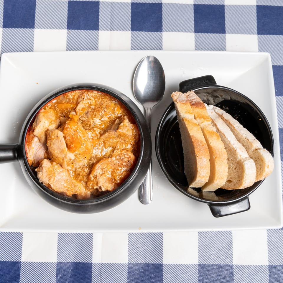Chicken stew in a black pot with sliced bread on a white plate and blue checkered tablecloth.