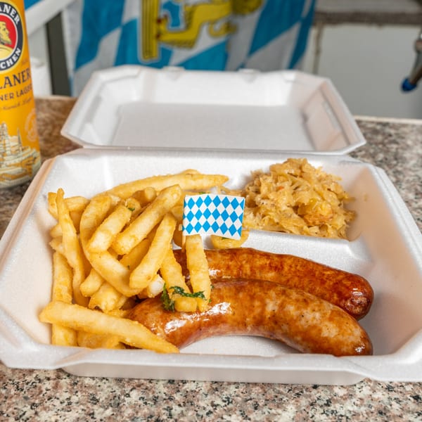 Bratwurst, fries, and sauerkraut in a to-go container with a Bavarian flag toothpick and beer can.