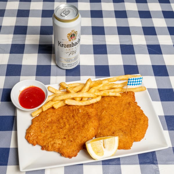 Two breaded schnitzels with fries, ketchup, lemon, and a Krombacher Pils beer can on a checkered tablecloth.