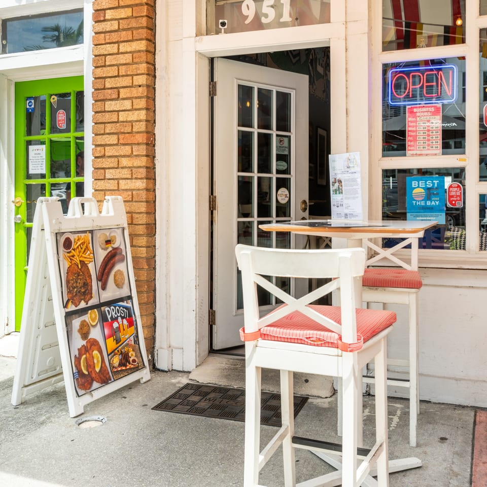 A restaurant storefront with an "OPEN" sign, outdoor seating, and a sandwich board displaying German food.